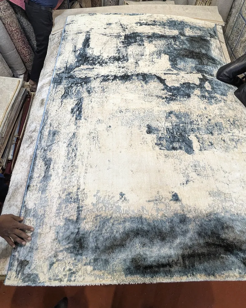 An abstract cream and blue-grey rug spread flat on a stack of rugs.