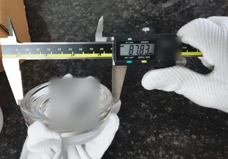 Gloved hands hold a clear glass jar between the jaws of a digital caliper reading 87.83 mm.