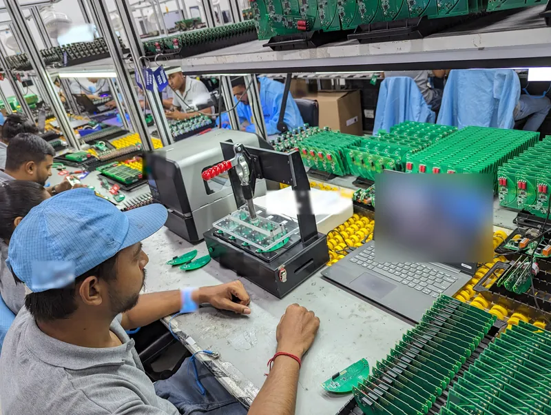 A technician seated beside a toggle-clamp test jig holding a green circuit board, with racks of tested boards around the bench.