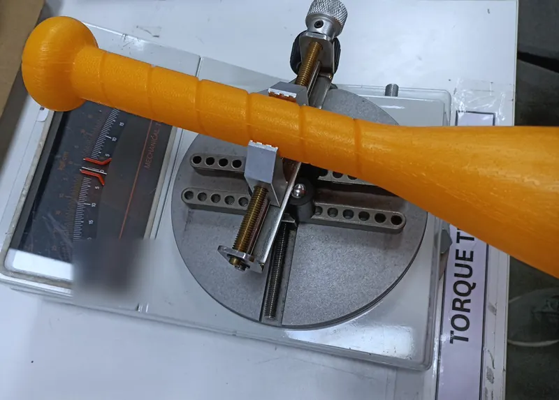 An orange moulded plastic bat clamped on a torque tester with an analogue scale.