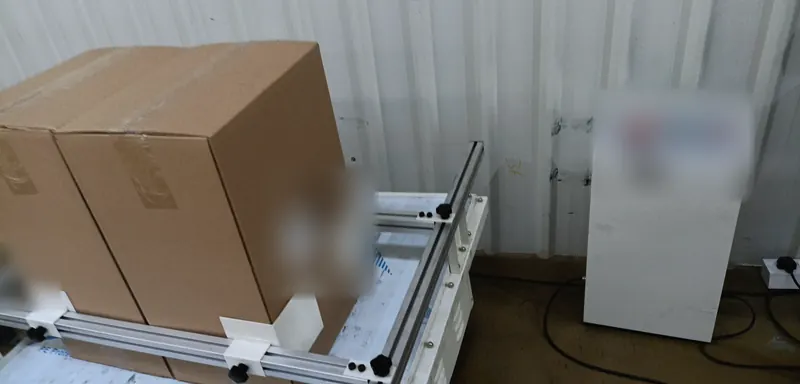 Plain export cartons clamped on the table of a transit vibration tester beside its control cabinet.