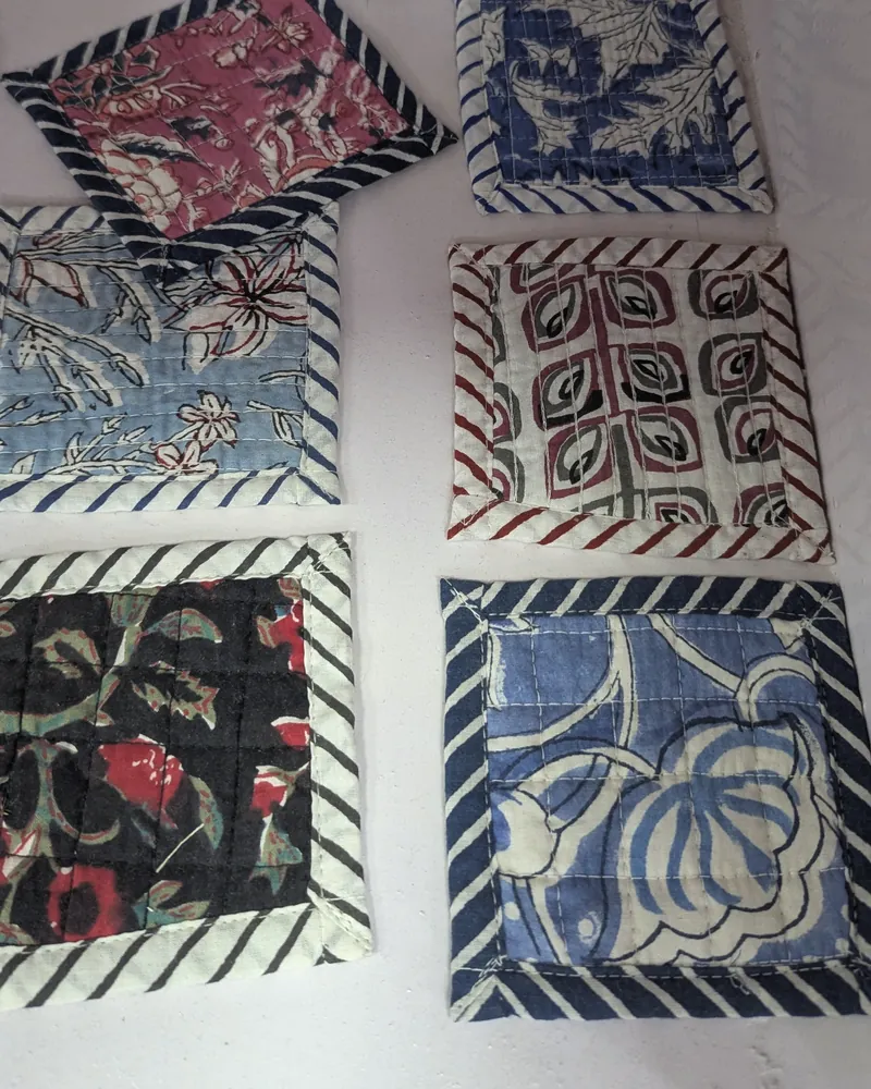Six quilted block-printed fabric coasters with striped borders laid on a white sheet.