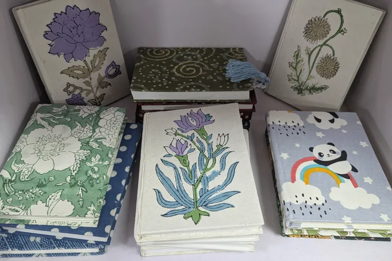 Hand block-printed fabric-covered notebooks with floral and panda designs, stacked on a shelf.