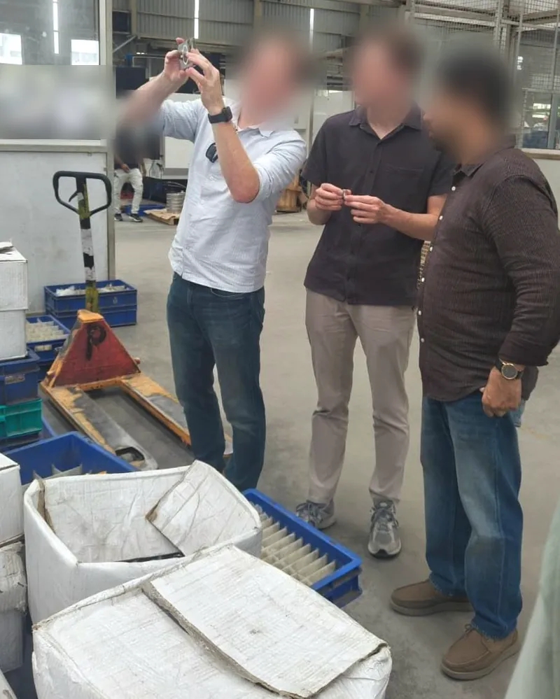 Three people on a factory floor, one holding a small metal part up to the light while the others look on; faces blurred.