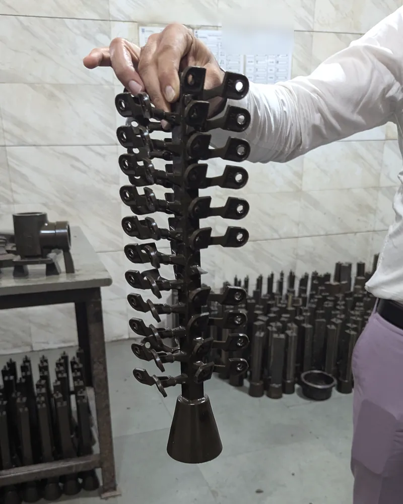 A hand holds up a dark wax tree of small bracket patterns on a central sprue, with more wax trees in the background.