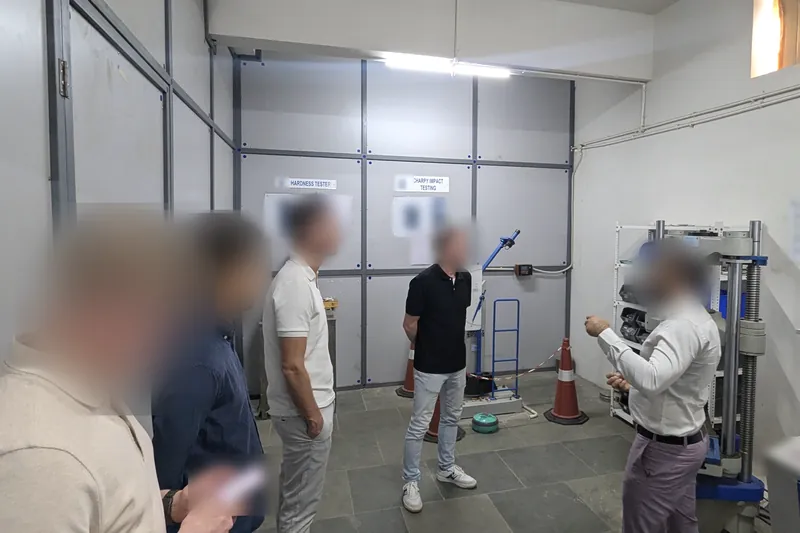 Visitors in a materials test room with a hardness tester, an impact tester and a universal testing machine; faces blurred.