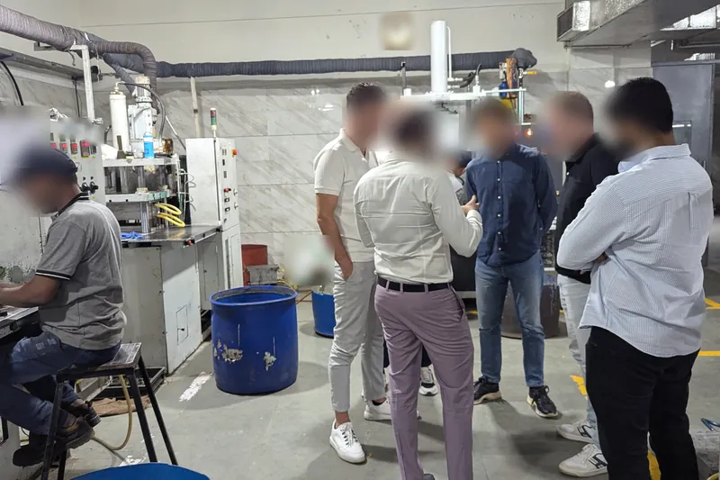 A group of visitors in conversation on a casting shop floor beside wax-injection presses and blue drums; all faces blurred.